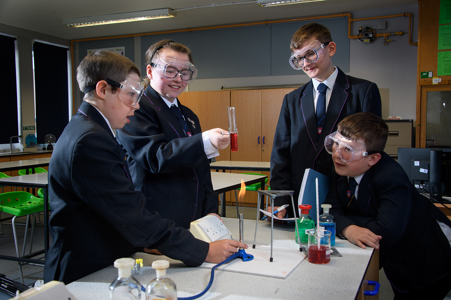 Four students, three male one female wearing goggles, holding a test tube over a bunsen burner flame