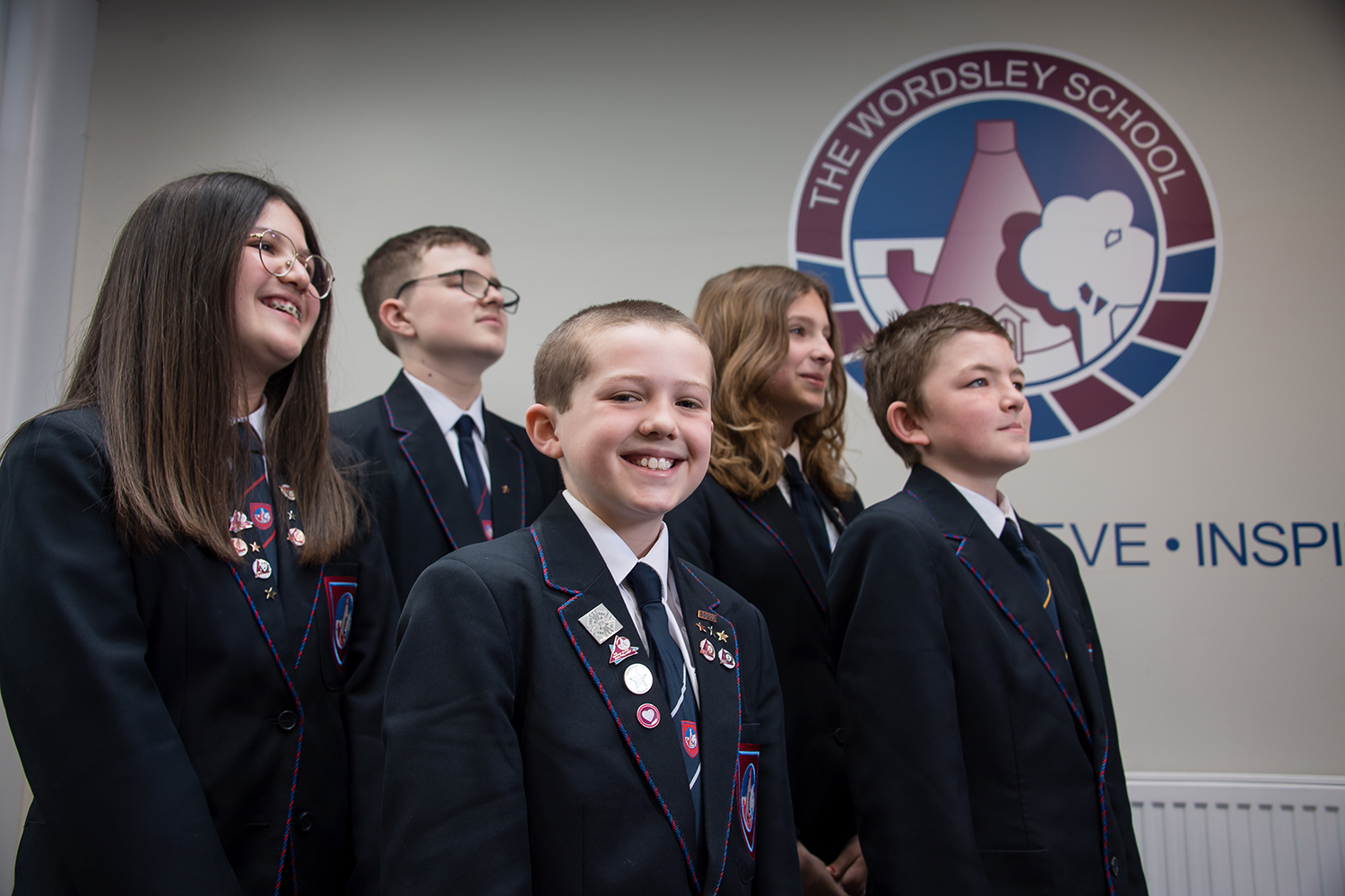 Five students in uniform standing in front of Wordsley logo on the wall. One boy at the front smiling at the camera