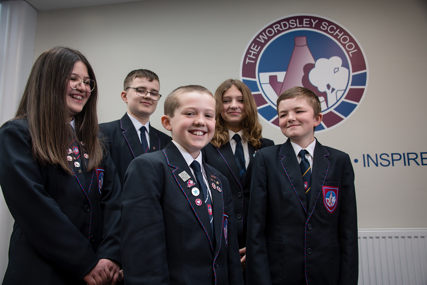 Five students in uniform standing in front of Wordsley School logo on the wall. Four students looking and smiling at boy at the front
