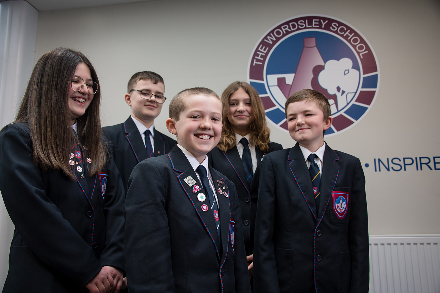 Five students in uniform standing in front of Wordsley School logo on the wall. Four students looking and smiling at boy at the front