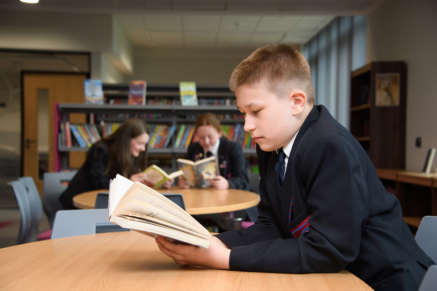 Boy reading a book in the library with two students reading in the background