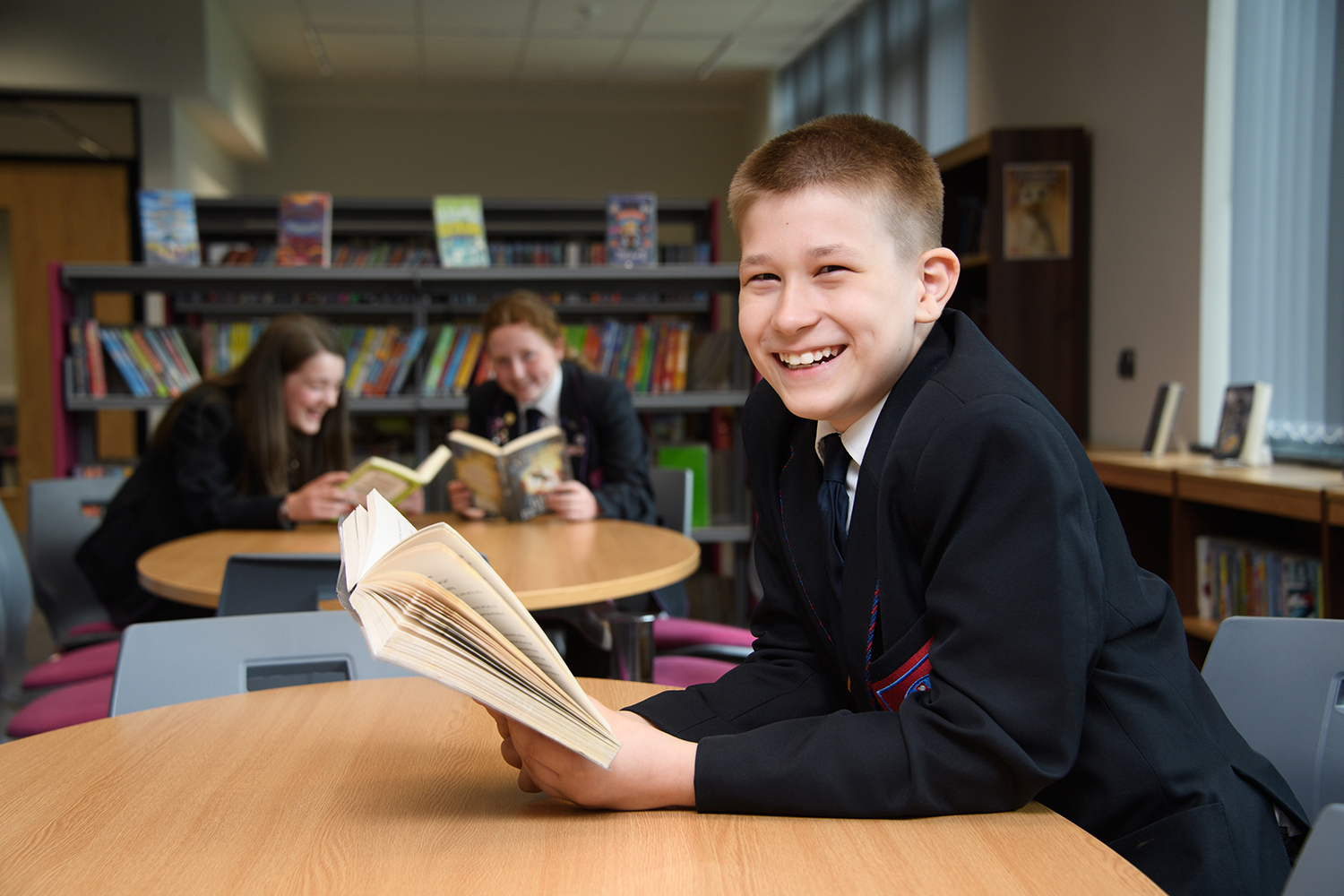 Boy in the library holding a book and smiling at the camera, with two students reading in the background