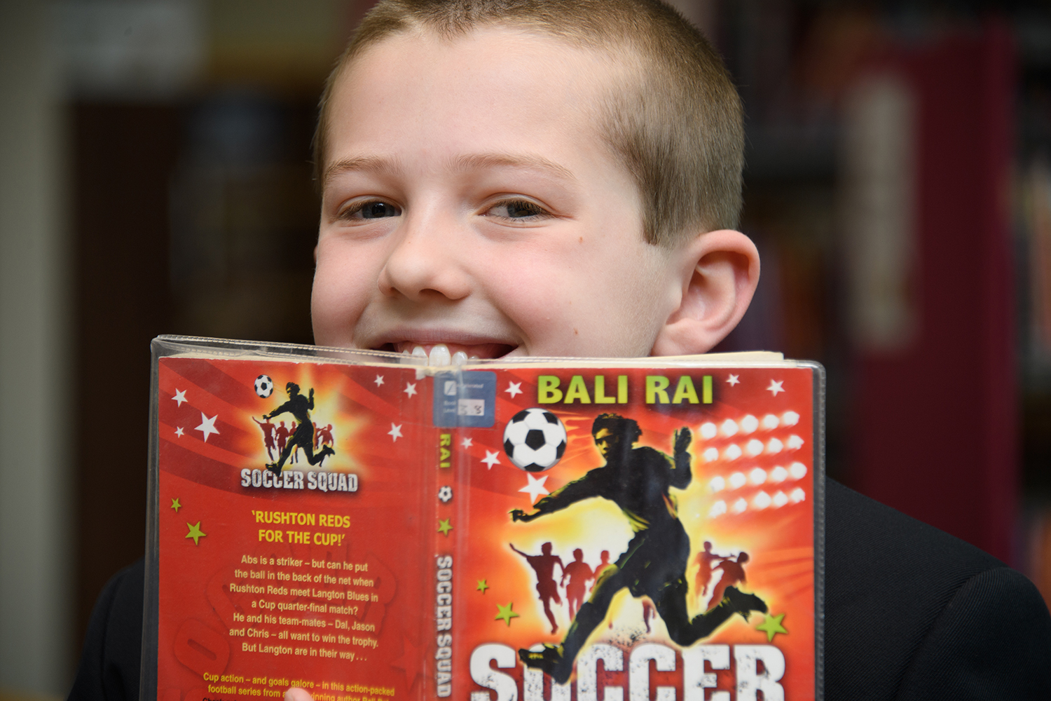 Boy in the library smiling over the top of a book