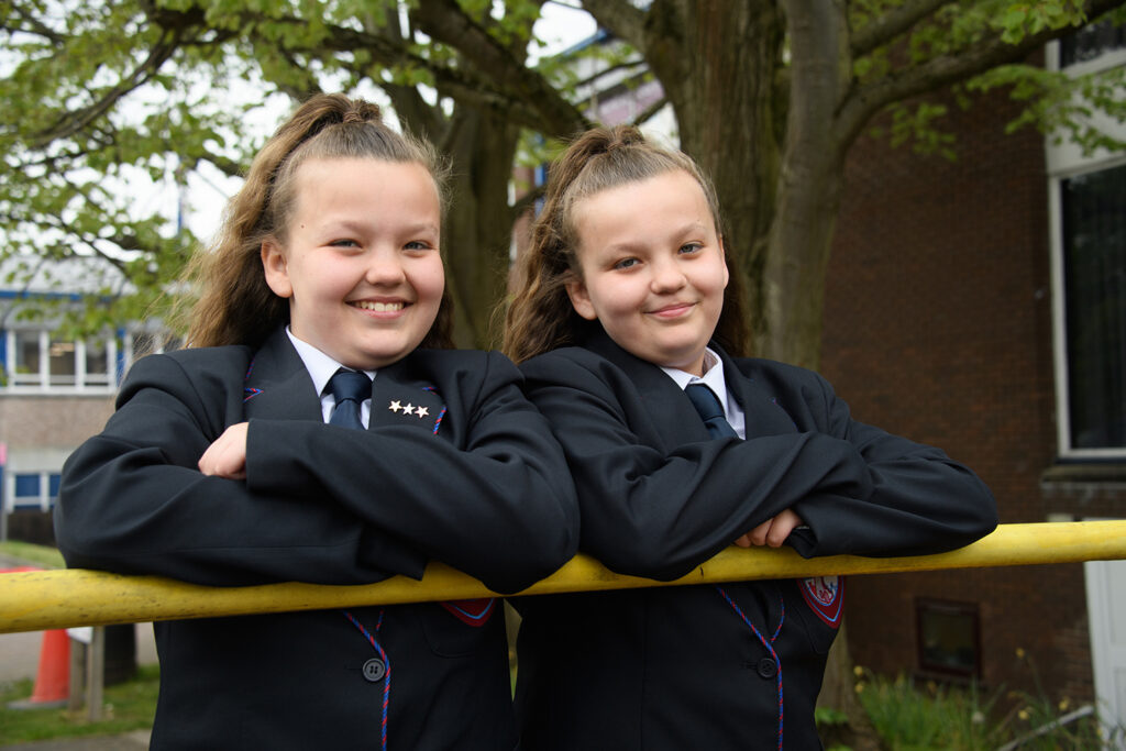 Twin girl students leaning on pole smiling at camera