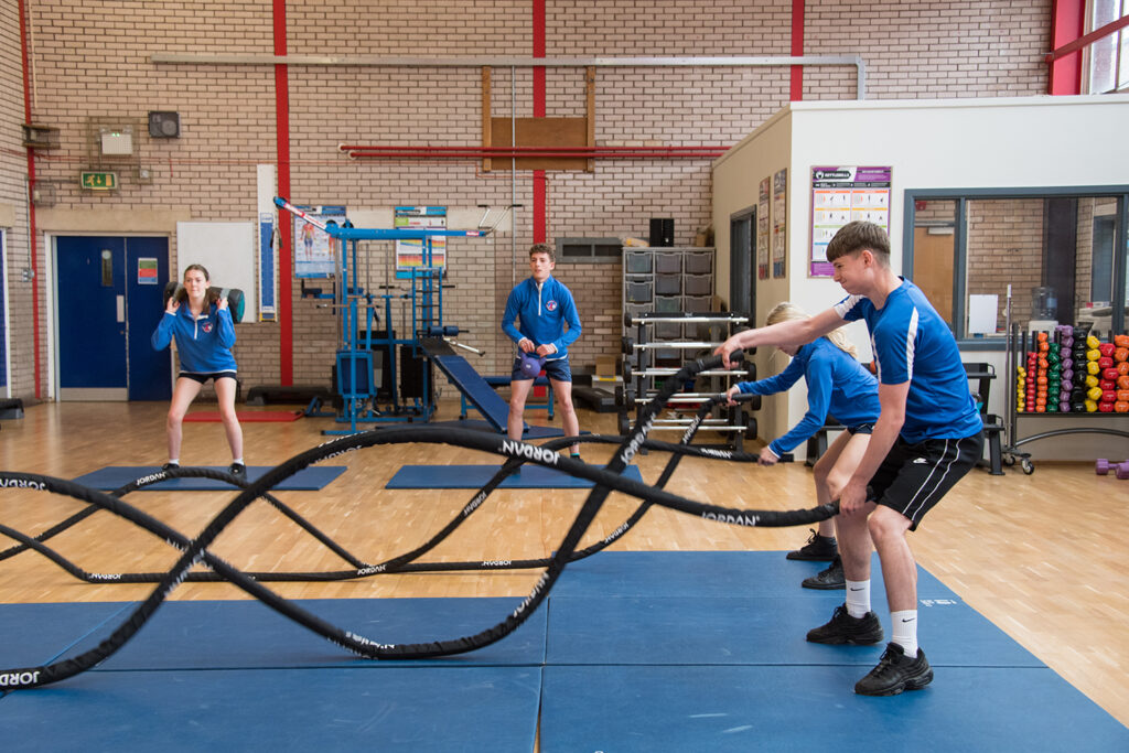 Male and female students in PE kits, two using weighted ropes and two lifting weights in sports hall