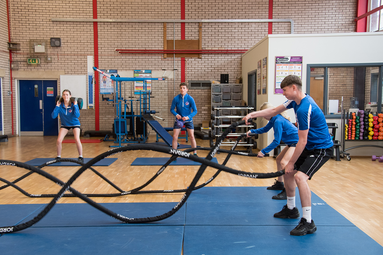 Male and female students in PE kits, two using weighted ropes and two lifting weights in sports hall