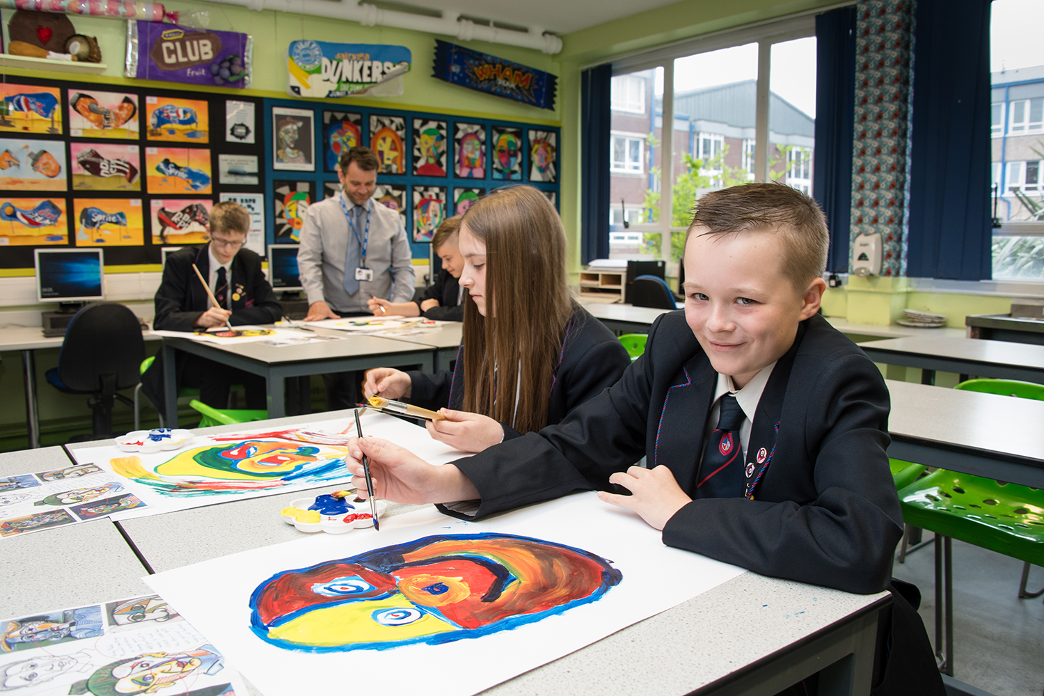 Two students painting in an art room smiling with a teacher helping students in the background