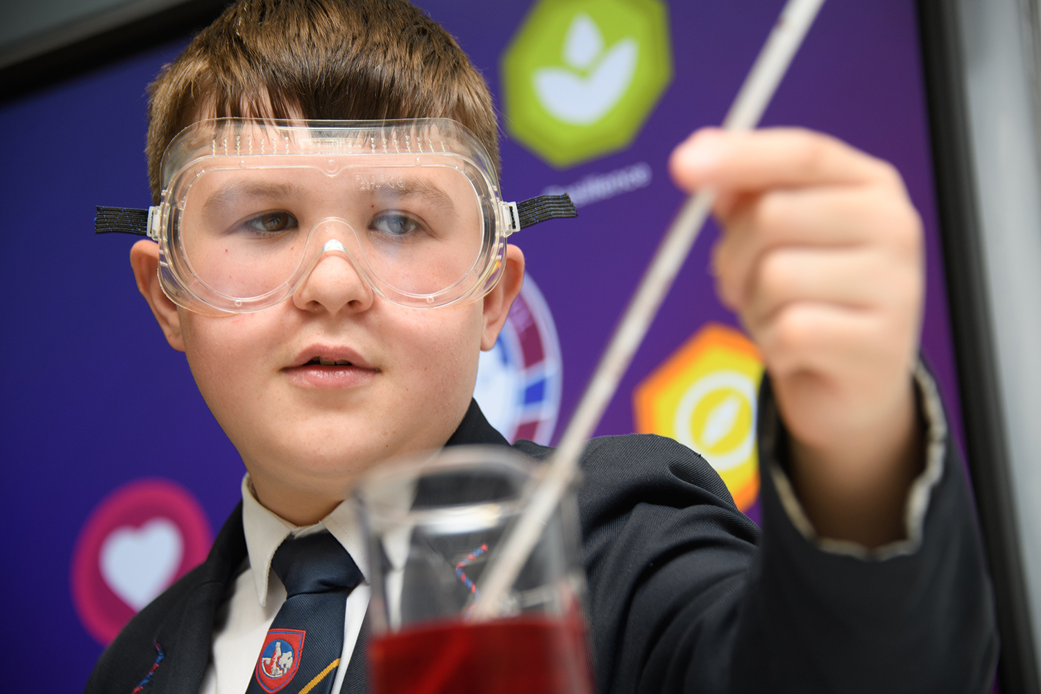 Boy in science room wearing goggles and holding a thermometer in a beaker of red liquid