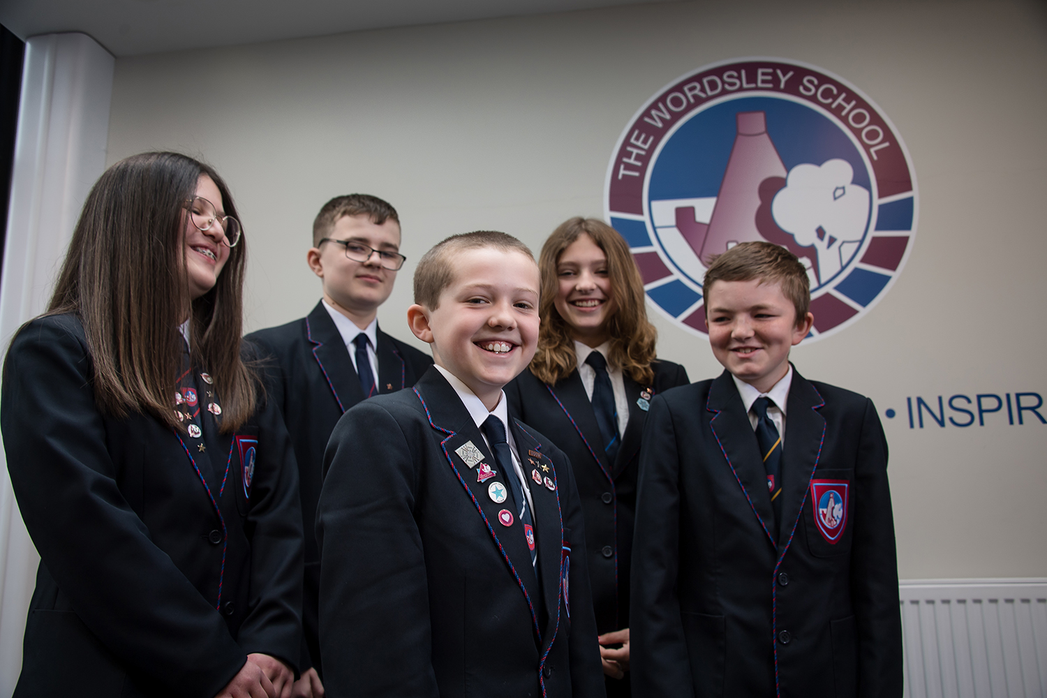 Five students in uniform standing in front of The Wordsley Logo on the wall, smiling into the distance