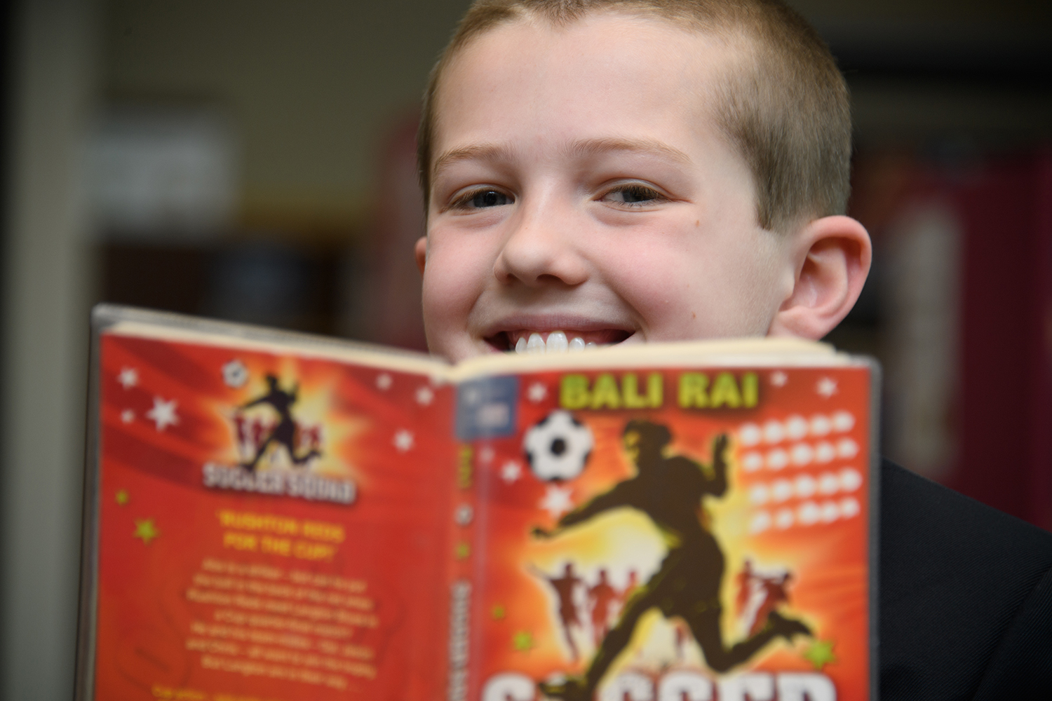 Boy in the library smiling over the top of a book