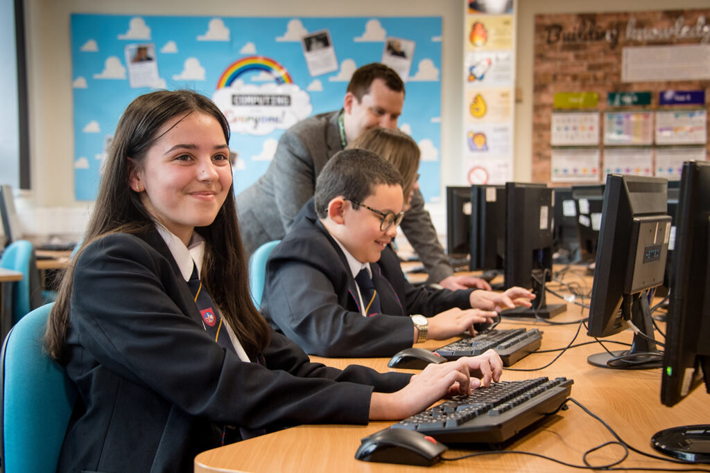 Three students sitting in computer room with teacher. Girl at front smiling at camera