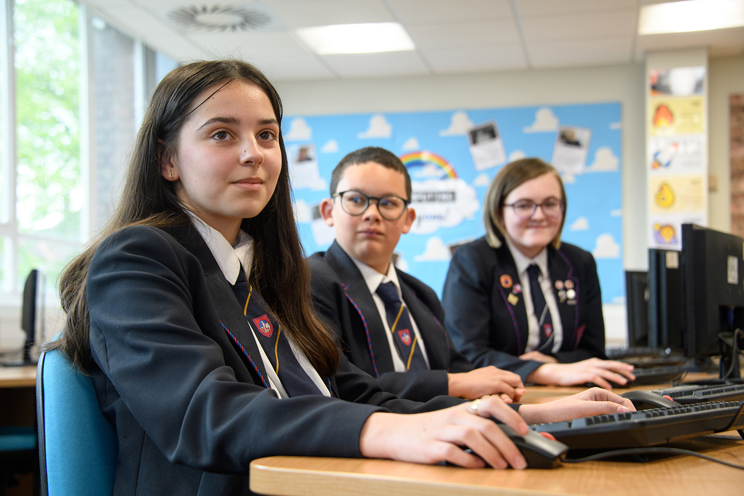 Three students sitting at computers, girl at the front smiling at the camera