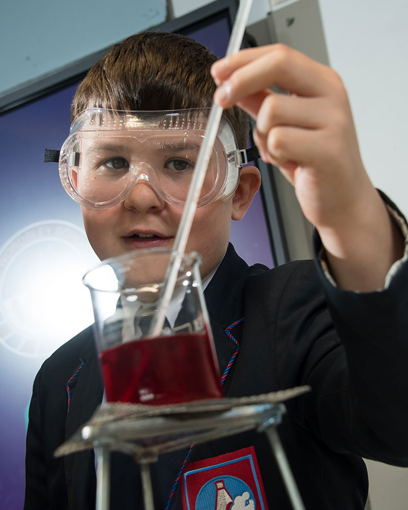 Boy wearing goggles holding thermometer in beaker of red liquid