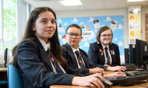 Three students sitting at computers, girl at the front smiling at the camera